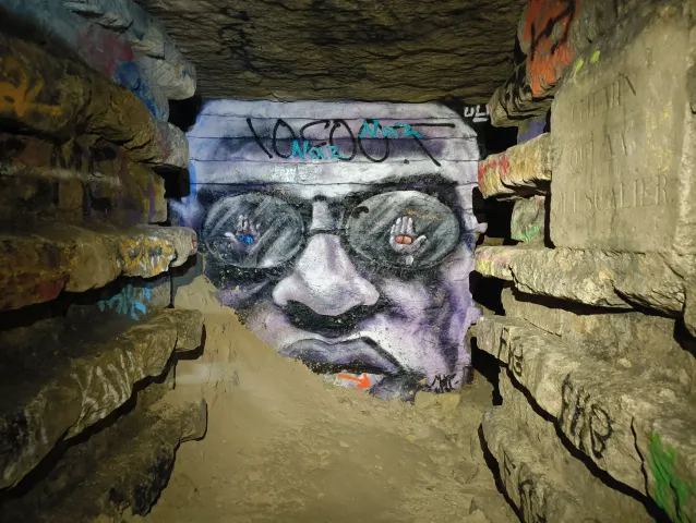 Visage à lunettes peint au fond d'une galerie basse, entre des banquettes de calcaire